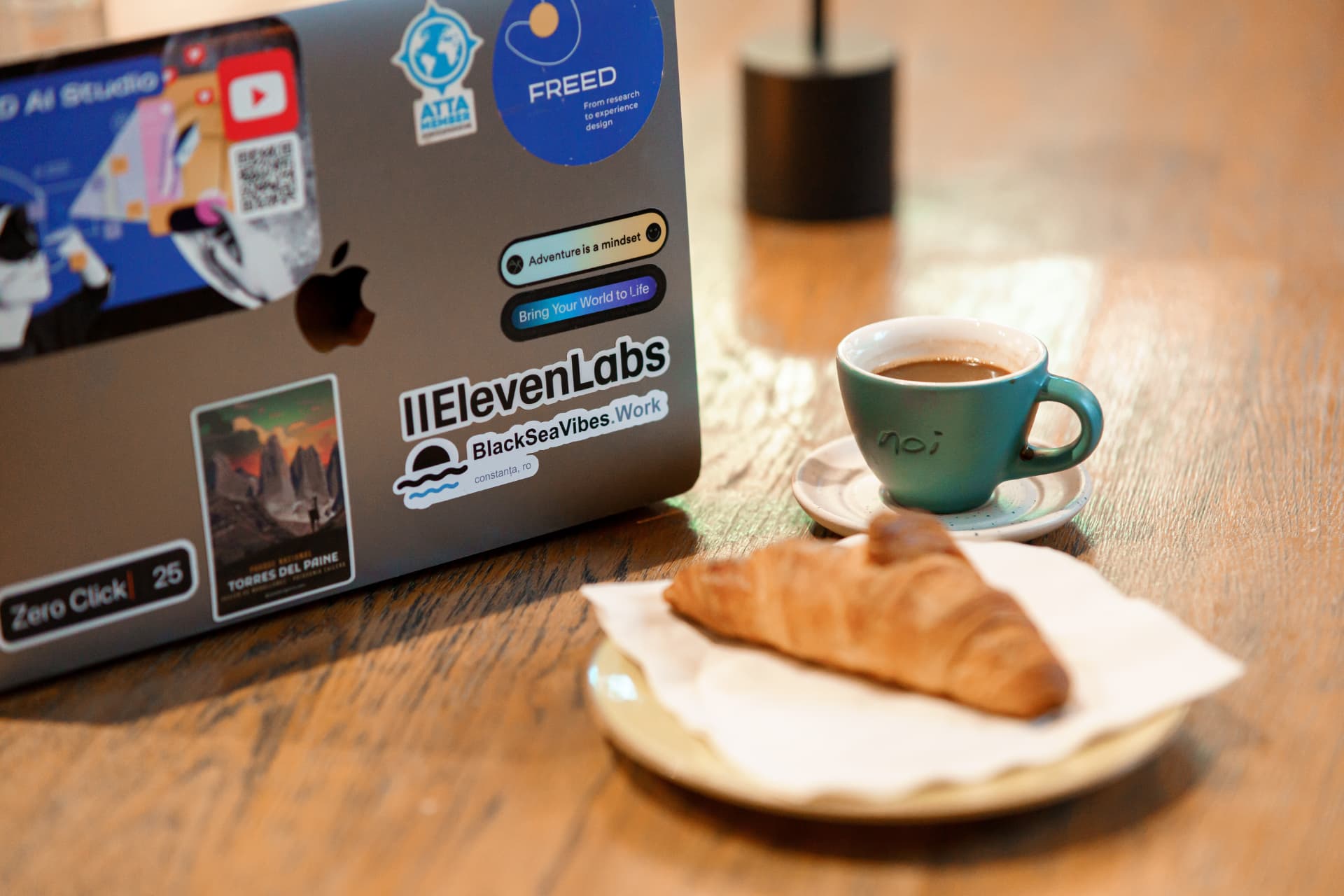 Computador portátil con adhesivo de ElevenLabs junto a una taza de café y un croissant